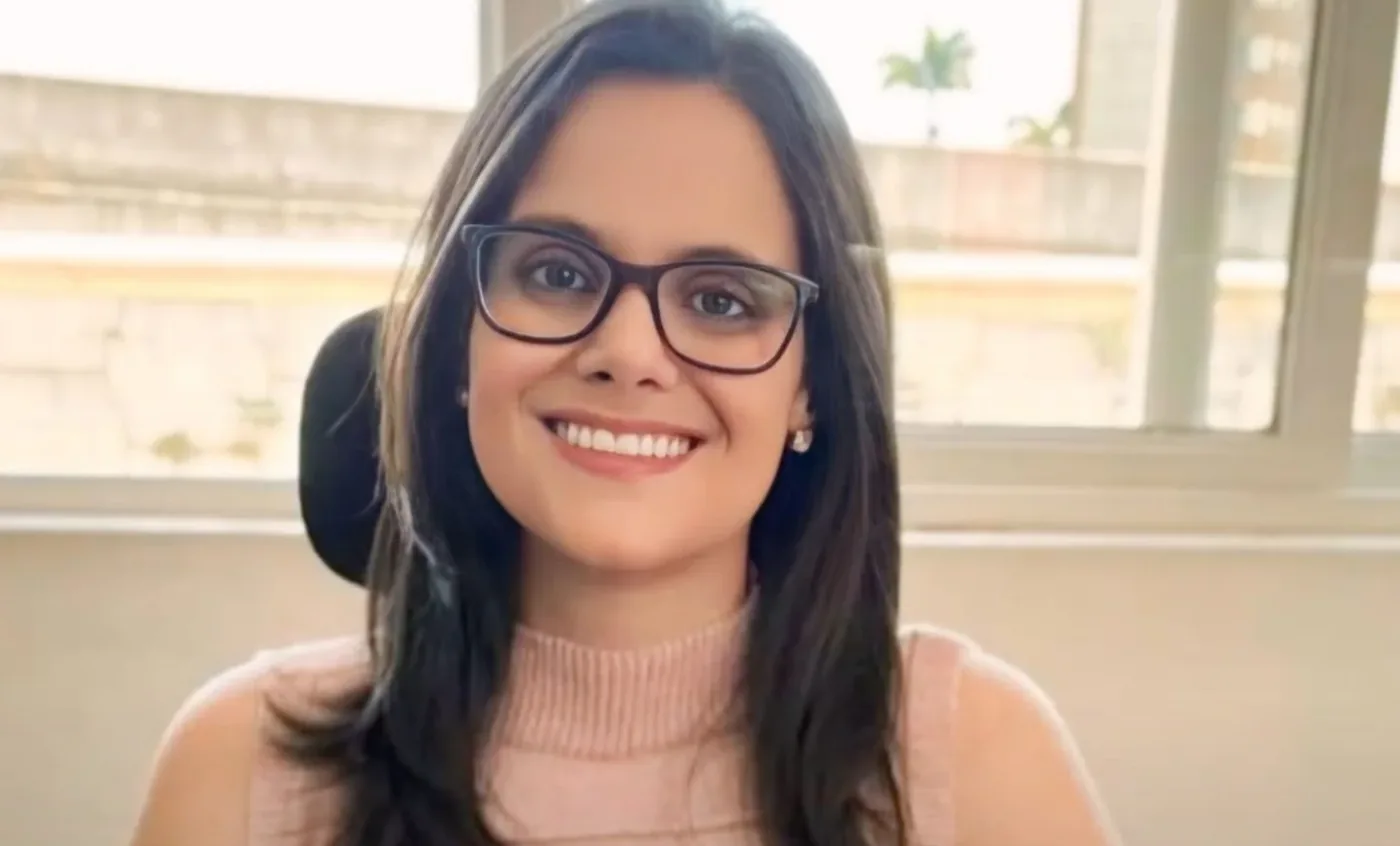 Marina Meirelles de óculos e blusa rosa, sorrindo, sentada à mesa